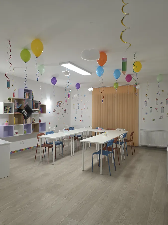 Sala de clasă luminoasă cu mese albe, scaune colorate și rafturi, decorată cu baloane