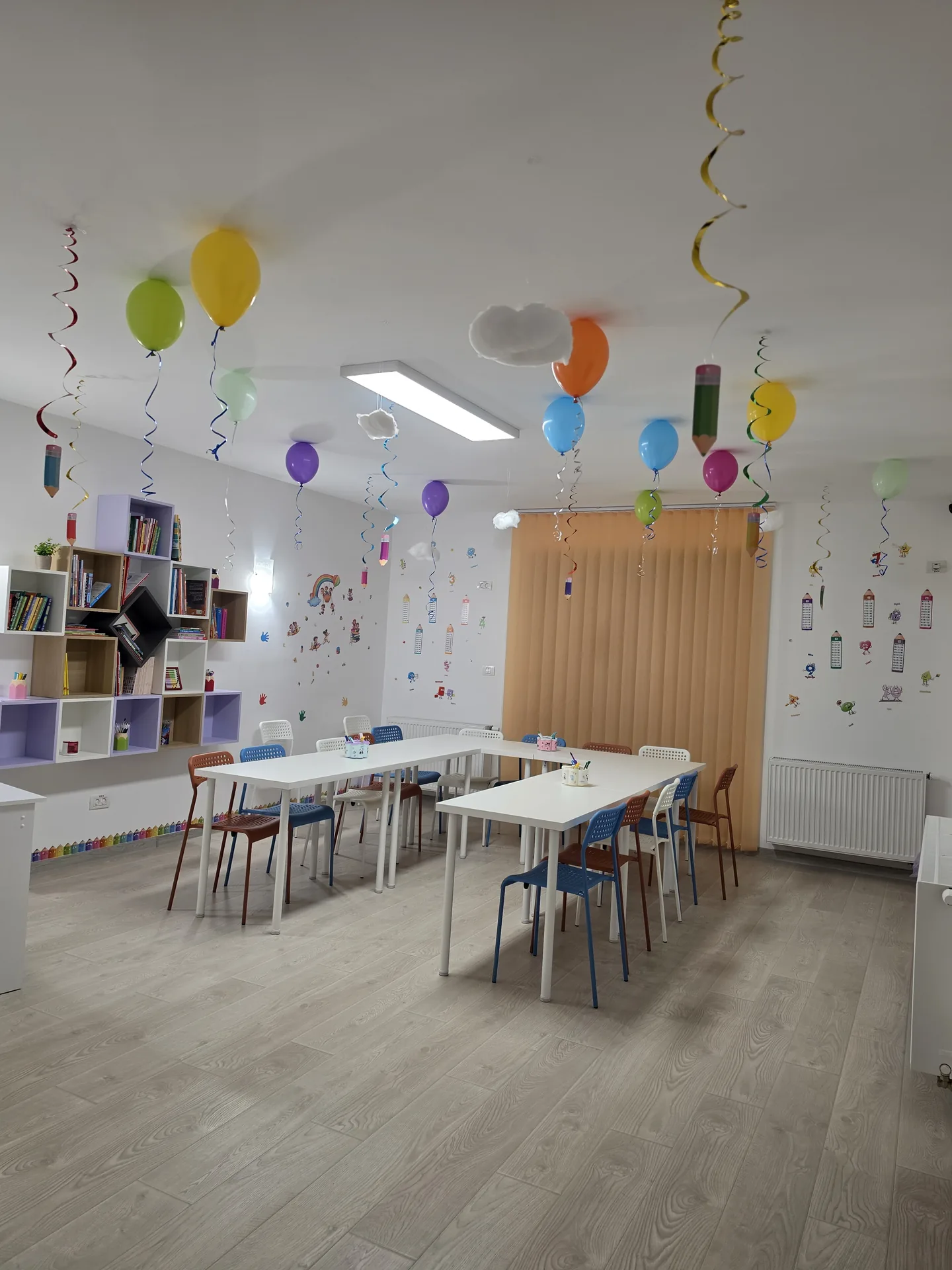 Sala de clasă cu mese de lucru, bibliotecă și perete decorat cu autocolante vesele