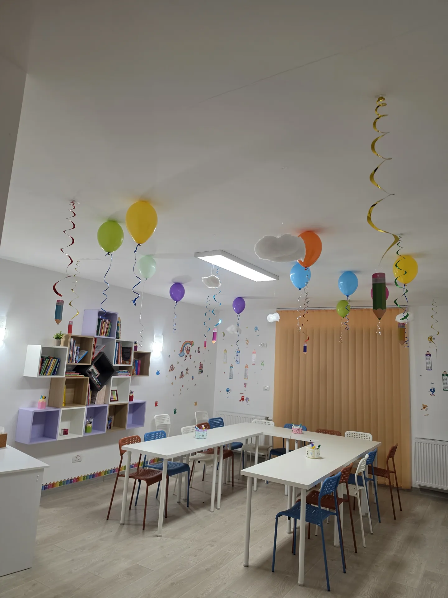 Sala de clasă cu mese albe, bibliotecă și decorațiuni colorate pe pereți